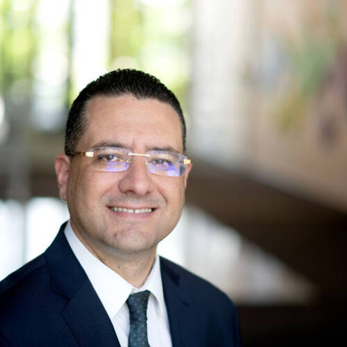 A person with short, dark, neatly styled hair is wearing a dark suit, white shirt, and a dark patterned tie, posed in a modern indoor setting with large windows and abstract wall art in the background.